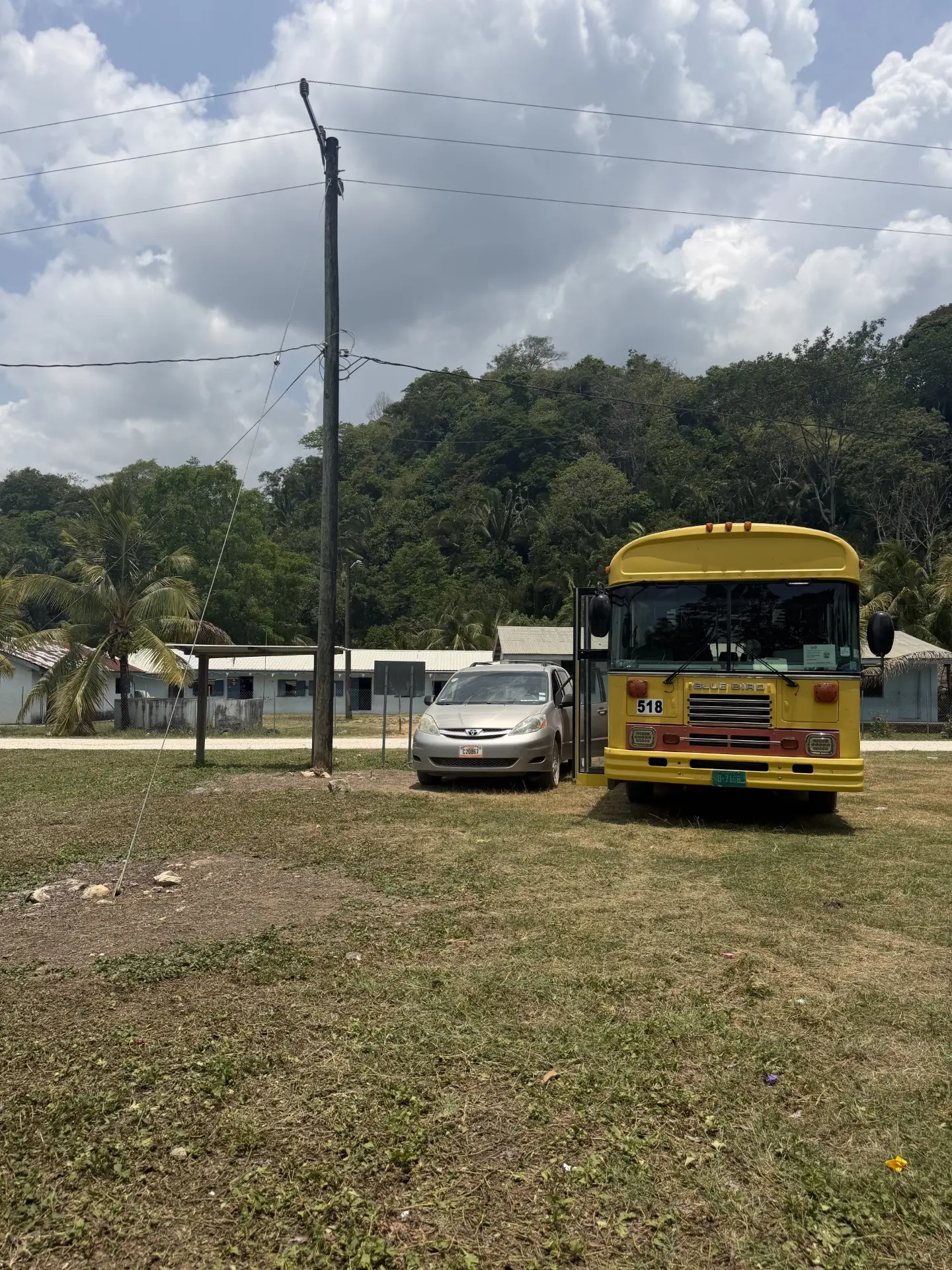 A yellow school bus parked on a grass field beside a small village school in rural Belize